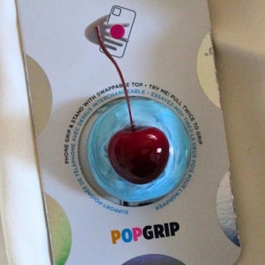 May include: A PopSockets PopGrip with a shiny, deep red cherry design. The cherry is set on a light blue base, and the packaging displays the PopSockets and PopGrip logos. The packaging also includes the text: "PHONE GRIP & STAND WITH SWAPPABLE TOP".