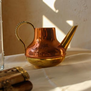 May include: A copper and brass watering can with a woven handle and a long spout. The watering can is on a white surface. The copper has a shiny finish, and the brass has a golden hue.