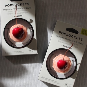 Könnte beinhalten: Zwei PopSockets Magnetic Phone Grip & Stand-Boxen. Jede Box zeigt ein Kirschdesign. Die Kirsche ist rot mit einem braunen Stiel, auf einer rosa Basis. Die Boxen sind weiß mit schwarzem Text und sind mit MagSafe-Ladegeräten kompatibel.