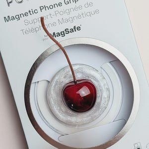 Puede incluir: Un soporte y agarre magnético para teléfono PopSockets en su empaque minorista. El producto presenta un diseño de cereza de color rojo oscuro brillante con un tallo marrón, centrado en una base circular blanca, translúcida y brillante.