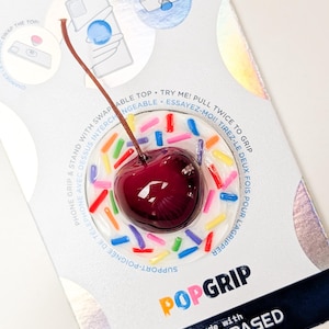 May include: A PopGrip phone accessory featuring a dark red cherry with a brown stem centered on a white circular base decorated with colorful sprinkles. The packaging includes the text PopSockets, PopGrip, and made with plant-based materials.
