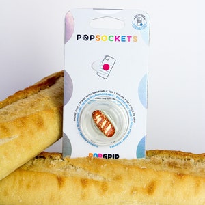 May include: A white PopSockets phone grip with a brown and white baguette design. The grip is on a white background with the PopSockets logo and the words "PopGrip" and "Support-Poignée de Téléphone" printed on the packaging.