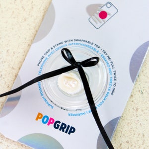 May include: A white PopSocket phone grip with a black ribbon tied around it. The grip has a clear plastic top with a white, pearl-like button. The packaging says "PopSocket" and "PopGrip" in colorful letters.