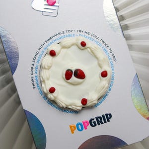 May include: A white PopSocket phone grip with a cake design, featuring red strawberry and cherry accents. The packaging includes the text "POPGRIP" and instructions in English and French.