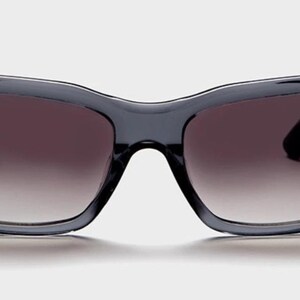 May include: Dark gray sunglasses with a gradient lens. The frames are a solid, rectangular shape with a slight curve at the top. The lenses transition from dark at the top to a lighter shade at the bottom. The sunglasses are set against a white background.