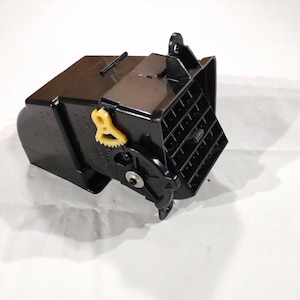 Op de afbeelding: Een zwarte kunststof auto-ventilatieopening met een gele tandwiel en een reeks verticale lamellen. De ventilatieopening heeft een rechthoekige vorm met een gebogen gedeelte aan één kant. De afbeelding is genomen tegen een witte achtergrond.