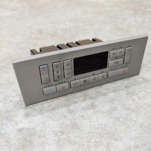 May include: A rectangular automotive climate control panel in a matte gray finish. The unit features a central black digital display screen surrounded by various push buttons for air conditioning, fan speed, temperature, and seat heating functions.