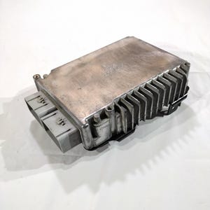 May include: A silver-colored electronic control module with a rectangular shape and a series of cooling fins on one side. Two gray connectors are visible on the left side. The module is likely for automotive or industrial use.