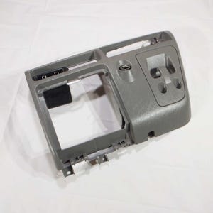 May include: A gray plastic car dashboard component with various openings and mounting points. The piece has a rectangular opening in the center, a small compartment, and several button or switch cutouts. The overall design is functional and automotive-focused.