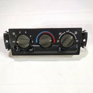 May include: A black car air conditioning control panel with three rotary dials and several buttons. The dials have textured grips and are labeled with numbers and symbols. The panel includes controls for fan speed, temperature, and air conditioning.