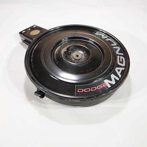 May include: A black air cleaner with the word "MAGNUM" in white and green lettering and the word "DODGE" in red lettering. The air cleaner is circular with a metal housing and a central mounting point.