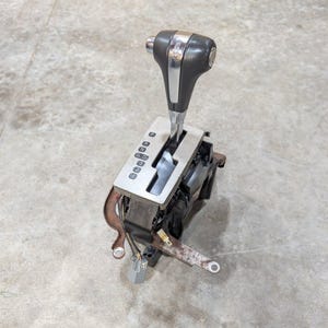 Op de afbeelding: Een donkergrijze en verchroomde automatische versnellingspook met een verticale schakelplaat met de markeringen P, R, N, D4, D3, 2 en 1. De unit bevat een metalen basis met montagebeugels en elektrische aansluitingen.