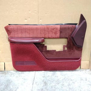Puede incluir: Panel de puerta de coche burdeos con una superficie afelpada y texturizada. El panel tiene forma rectangular con un reposabrazos integrado y una carcasa para altavoz. El diseño incluye una sección de tela estampada y ranuras de ventilación horizontales.