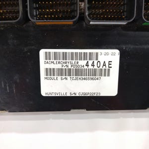 May include: A black electronic control module with multiple connectors. A white label displays "DAIMLERCHRYSLER" and "440AE" along with other text and barcodes. The module is likely for automotive use.