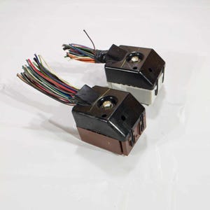 Puede incluir: Dos conectores eléctricos negros con varios cables conectados. Los conectores tienen una base de plástico marrón y una parte superior de plástico negro. Los cables son de varios colores, incluyendo rojo, azul, verde, amarillo y blanco.