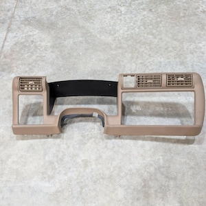 May include: A tan automotive dashboard bezel featuring a central black panel, integrated air conditioning vents on the left and right sides, and rectangular openings for instrument clusters and controls, constructed from molded plastic.