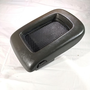 May include: A dark gray plastic car console cup holder with a textured black rubber insert. The cup holder has a rectangular shape with rounded corners and a small button on the side.