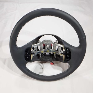 Può includere: Un volante grigio scuro con un bordo rivestito in pelle e cuciture visibili. Il volante ha pulsanti di controllo integrati e un telaio in metallo con cablaggio e connettori. Il design è circolare, incentrato sulla funzionalità e sull'estetica automobilistica.