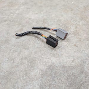 May include: Two electrical wire connectors with multi-colored wires wrapped in black electrical tape. One connector is black plastic with a rectangular shape, and the other is a light gray plastic connector with a similar rectangular design.