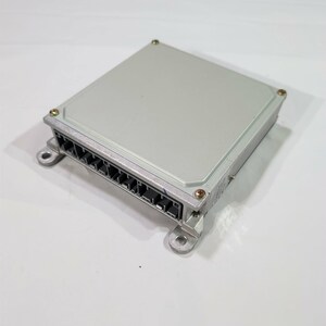 May include: A rectangular automotive electronic control unit with a light gray metal casing. The device features a black plastic connector port with multiple pin slots along one side and four metal mounting tabs with holes at each corner.