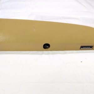 May include: A tan-coloured car component featuring a black circular aperture and a rectangular "DODGE" badge. The part has a curved form and seems to be made of plastic. The background is white.