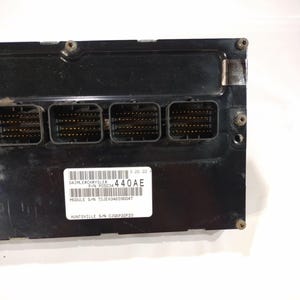May include: A black electronic control module with four connector ports. A white label displays text including "DAIMLERCHRYSLER", "440AE", and "MODULE S/N TCJEH346596047". The module has several screws and a metallic plate.