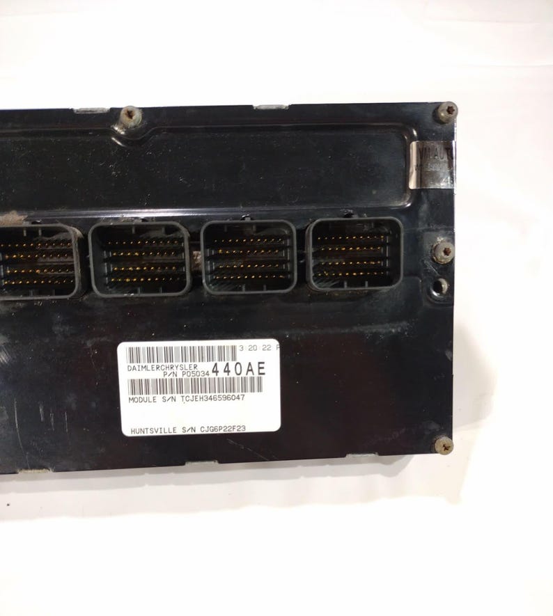 May include: A black electronic control module with four connector ports. A white label displays text including "DAIMLERCHRYSLER", "440AE", and "MODULE S/N TCJEH346596047". The module has several screws and a metallic plate.