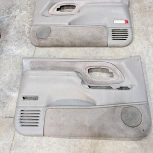 Puede incluir: Dos paneles de puerta interiores para automóvil en color beige, con rejillas de altavoz integradas, ranuras de ventilación horizontales y huecos para manijas. Los paneles combinan plástico liso con inserciones de tela texturizada.