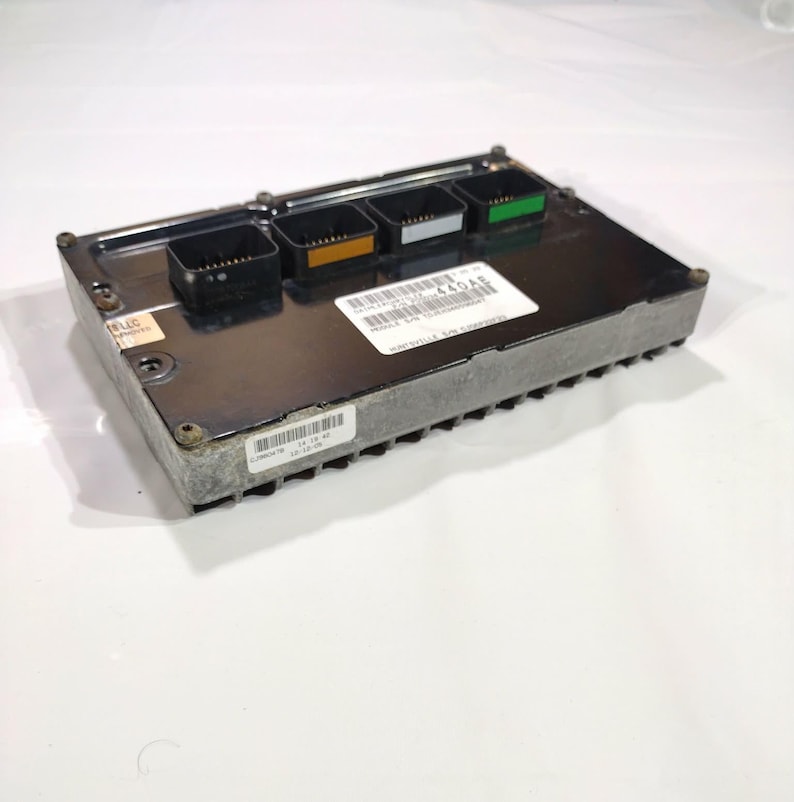 May include: A rectangular, silver electronic control module with four black connectors, each with a different coloured accent. A white label with black text and a barcode is affixed to the top. The module is on a white background.