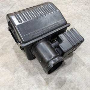 Pode incluir: Uma carcaça de filtro de ar automotivo de plástico preto com um corpo principal retangular, seções superiores nervuradas e uma porta de admissão cilíndrica. A carcaça tem gravado o texto: REPLACE WITH AC AIR FILTER A1200C USE A15010 FOR SEVERE DUST CONDITIONS.