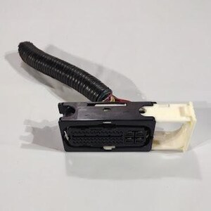 Op de afbeelding: Een zwarte elektrische connector met een beige beugel en een zwarte gegolfde kabel. De connector heeft meerdere pinnen en wordt waarschijnlijk gebruikt in auto- of elektronische toepassingen.