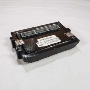 May include: A black rectangular automotive computer module with three grey connectors on top. The module has a label with text and barcodes. The module is on a white surface.