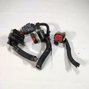 Puede incluir: Un surtido de mazos de cables eléctricos negros con varios conectores. Los conectores son negros, rojos y beige. El cableado está encerrado en una funda de plástico negro corrugado. Los extremos de algunos cables están expuestos.