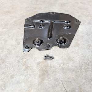 May include: A black metal mounting plate with a T-shaped raised ridge, multiple circular bolt holes, and two integrated threaded studs. Two separate small black hex bolts are positioned directly below the plate on the surface.