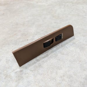 May include: A brown automotive interior door panel trim piece featuring a black power window switch and a black door lock switch. The door lock switch is clearly labeled with the text DOOR LOCK printed in white lettering on the brown surface.
