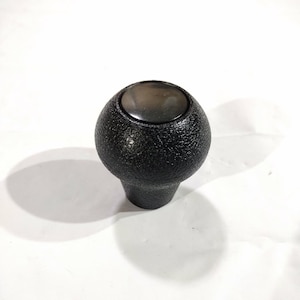 May include: A black, textured shift knob with a smooth, dark gray top. The knob has a rounded shape with a textured surface and a cylindrical base. The image is set against a white background.