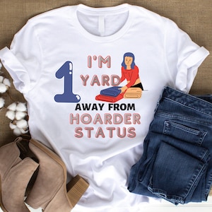 May include: White t-shirt with a blue number one and the text "I'm 1 yard away from hoarder status". The text is in pink and red. A cartoon illustration of a person in a red shirt and blue jeans is folding a stack of blankets.