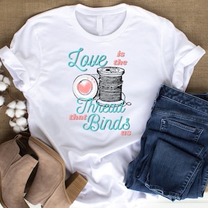 May include: White t-shirt with a teal and pink graphic of a spool of thread and the text "Love is the thread that binds us".