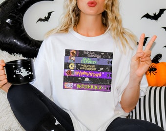 May include: A white t-shirt with a stack of VHS tapes featuring Halloween movie titles, including "Beetlejuice", "Halloweentown", "The Nightmare Before Christmas", "Little Monsters", and "Hocus Pocus".