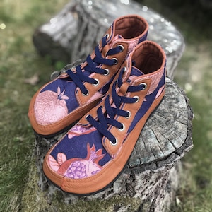 May include: A pair of high-top sneakers featuring a dark blue fabric upper with a pink pomegranate print. The shoes have tan leather trim, silver metal eyelets, and dark blue laces. The design includes a rounded toe box and a flat sole construction.