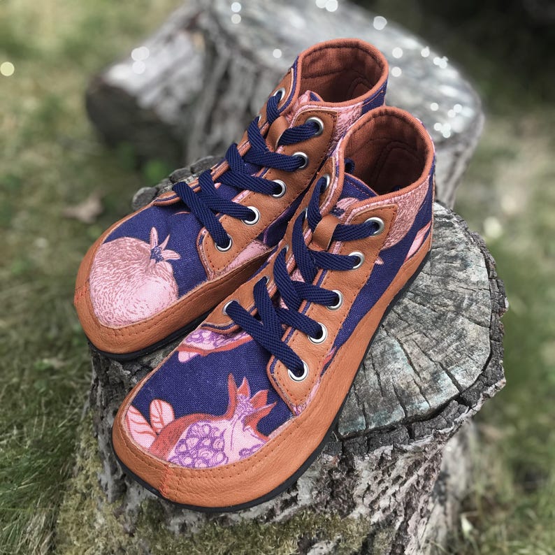 May include: A pair of high-top sneakers featuring a dark blue fabric upper with a pink pomegranate print. The shoes have tan leather trim, silver metal eyelets, and dark blue laces. The design includes a rounded toe box and a flat sole construction.