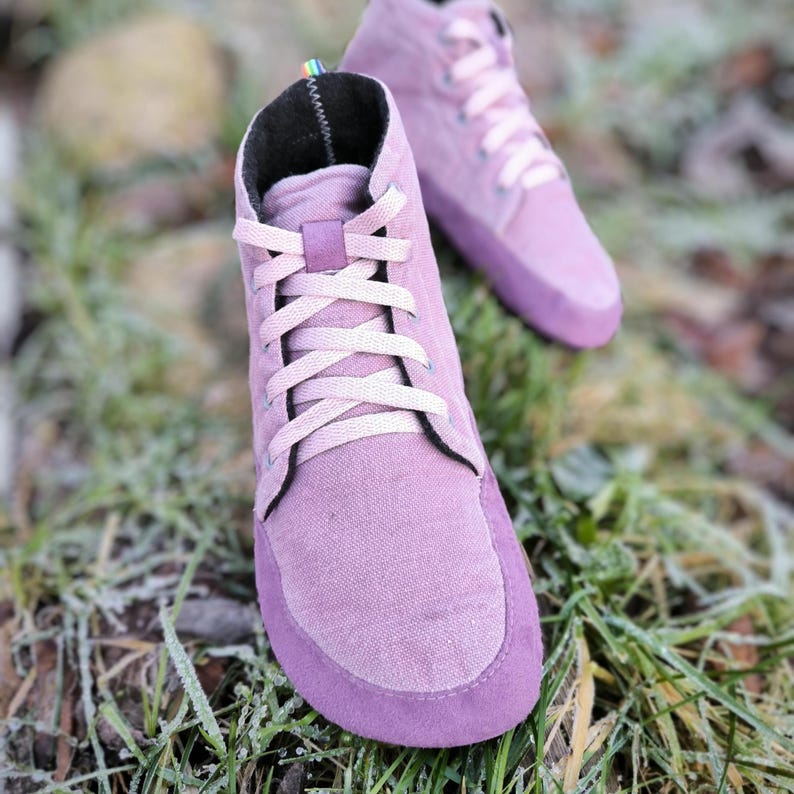 May include: A pair of high-top sneakers in a muted mauve color, featuring a canvas upper with a suede-like toe cap and heel reinforcement. The shoes have light pink laces, a black interior lining, and a small rainbow tab at the top of the heel.