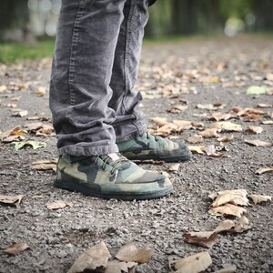 May include: A pair of low-top sneakers featuring a green, brown, and black camouflage pattern with black soles and laces, worn with dark gray corduroy trousers. The footwear has a rounded toe design and a small light-colored tag on the tongue.