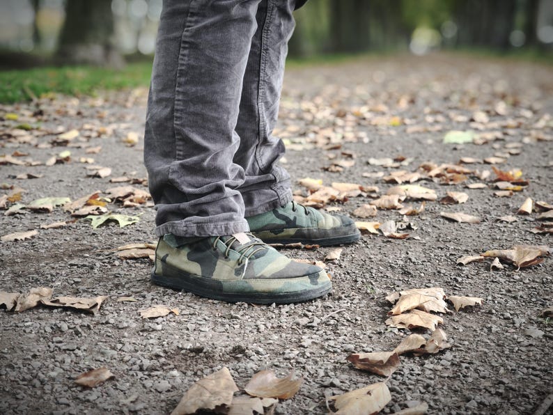 May include: A pair of low-top sneakers featuring a green, brown, and black camouflage pattern with black soles and laces, worn with dark gray corduroy trousers. The footwear has a rounded toe design and a small light-colored tag on the tongue.