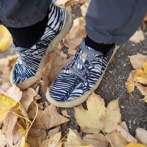May include: A pair of low-top sneakers featuring a black and white abstract leaf pattern on the upper fabric, grey laces, and a tan rubber sole. The footwear is worn with black socks and grey trousers, positioned among fallen yellow autumn leaves.