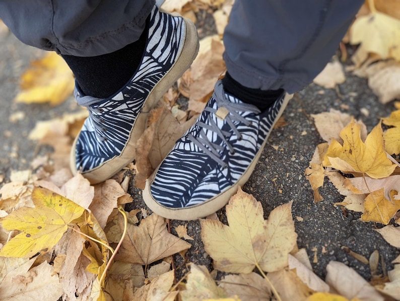 May include: A pair of low-top sneakers featuring a black and white abstract leaf pattern on the upper fabric, grey laces, and a tan rubber sole. The footwear is worn with black socks and grey trousers, positioned among fallen yellow autumn leaves.