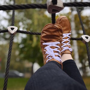 May include: A pair of tan leather sneakers featuring white laces, a rounded toe box, and a low-top silhouette. The shoes are worn with dark-colored pants and are positioned against a black rope climbing net structure with silver metal connectors.