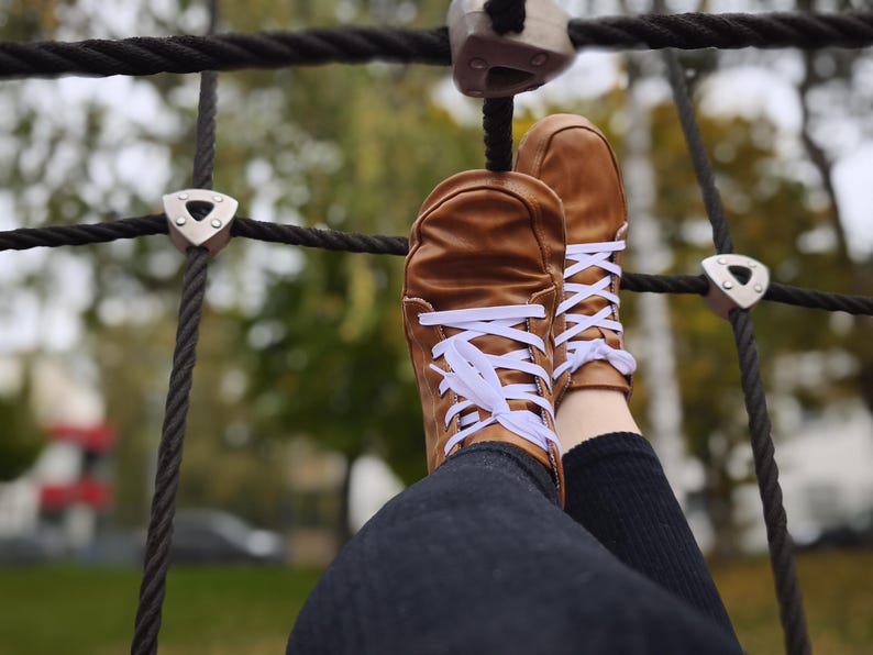 May include: A pair of tan leather sneakers featuring white laces, a rounded toe box, and a low-top silhouette. The shoes are worn with dark-colored pants and are positioned against a black rope climbing net structure with silver metal connectors.