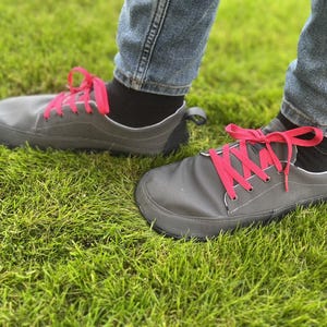 May include: A pair of low-top sneakers in a dark gray fabric with bright pink laces. The shoes feature a wide, rounded toe box, a black heel tab, and a thin, flexible black sole. The sneakers are worn with black socks and blue denim jeans.