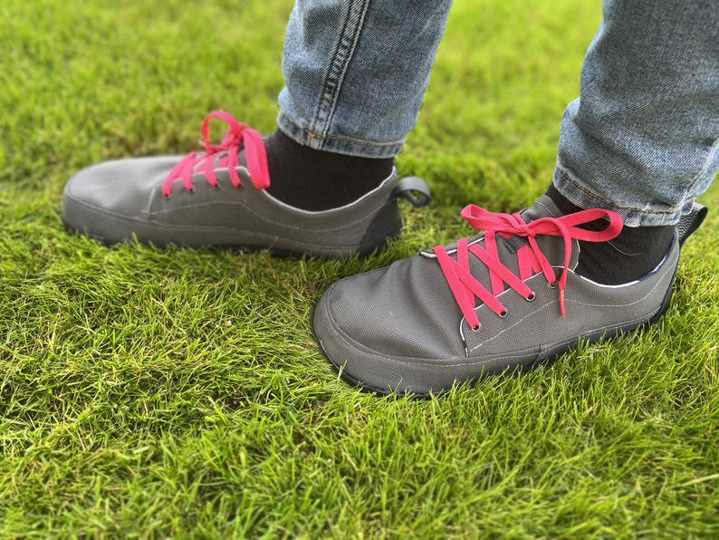 May include: A pair of low-top sneakers in a dark gray fabric with bright pink laces. The shoes feature a wide, rounded toe box, a black heel tab, and a thin, flexible black sole. The sneakers are worn with black socks and blue denim jeans.